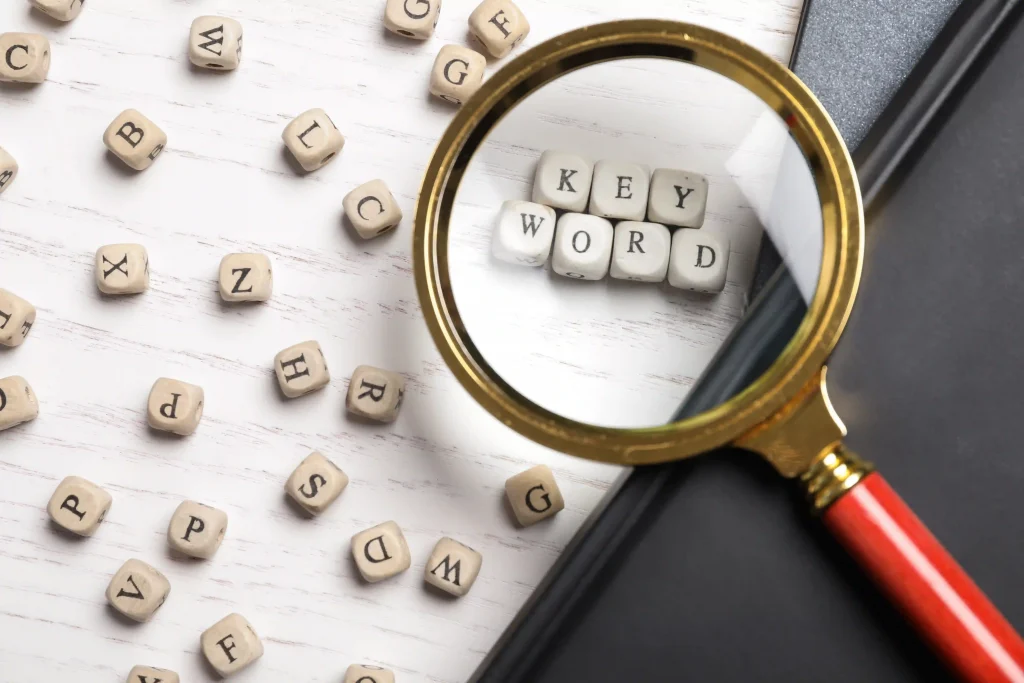 Magnifying glass with red shank over white dice forming the word keyword. Several other dice with letters on white table.