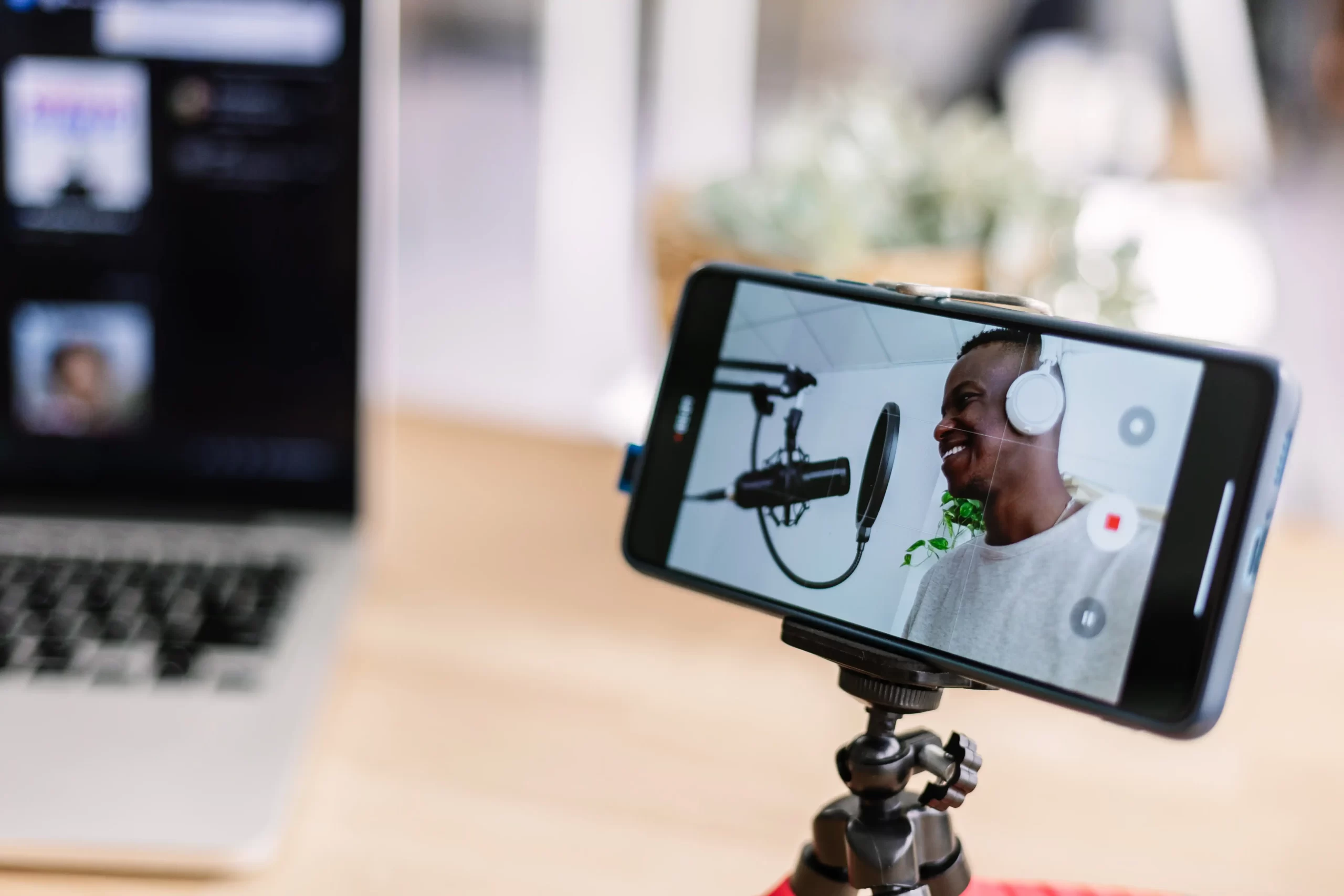 Smiling young man recording podcast with a smartphone mounted on a tripod, while streaming online at home studio.