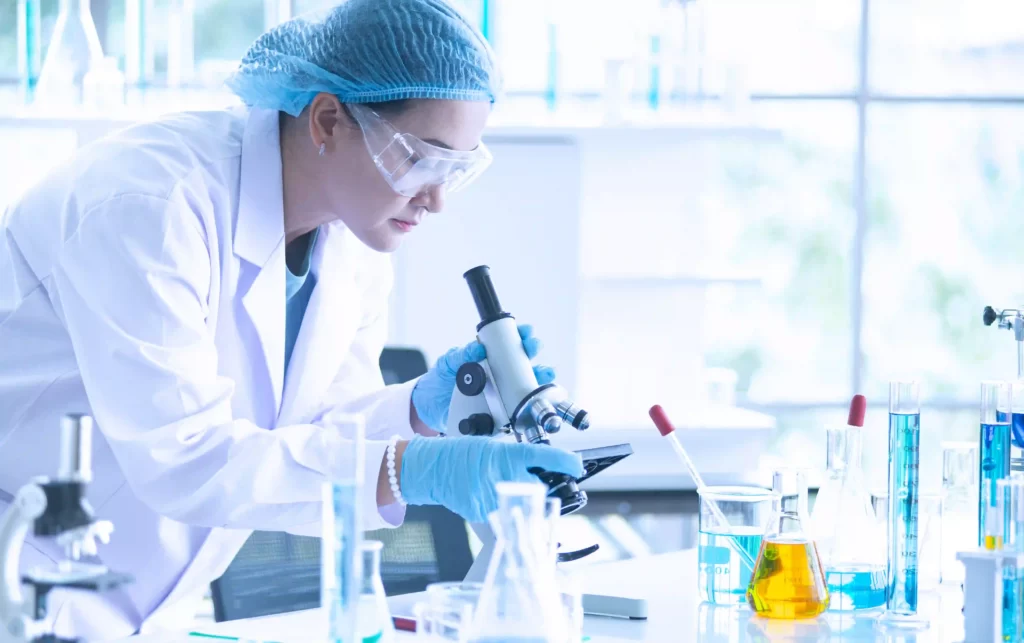 A female life-science scientists in a laboratory setting, dressed in a medical gown and safety glasses, working with microscope and liquid substances in various labware.