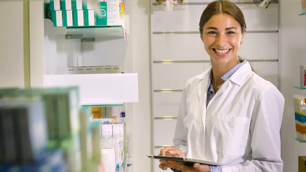 A female life-science scientist or a pharmacist, in a medical gown, standing in front of a shelf with various medical or pharmaceutical items.