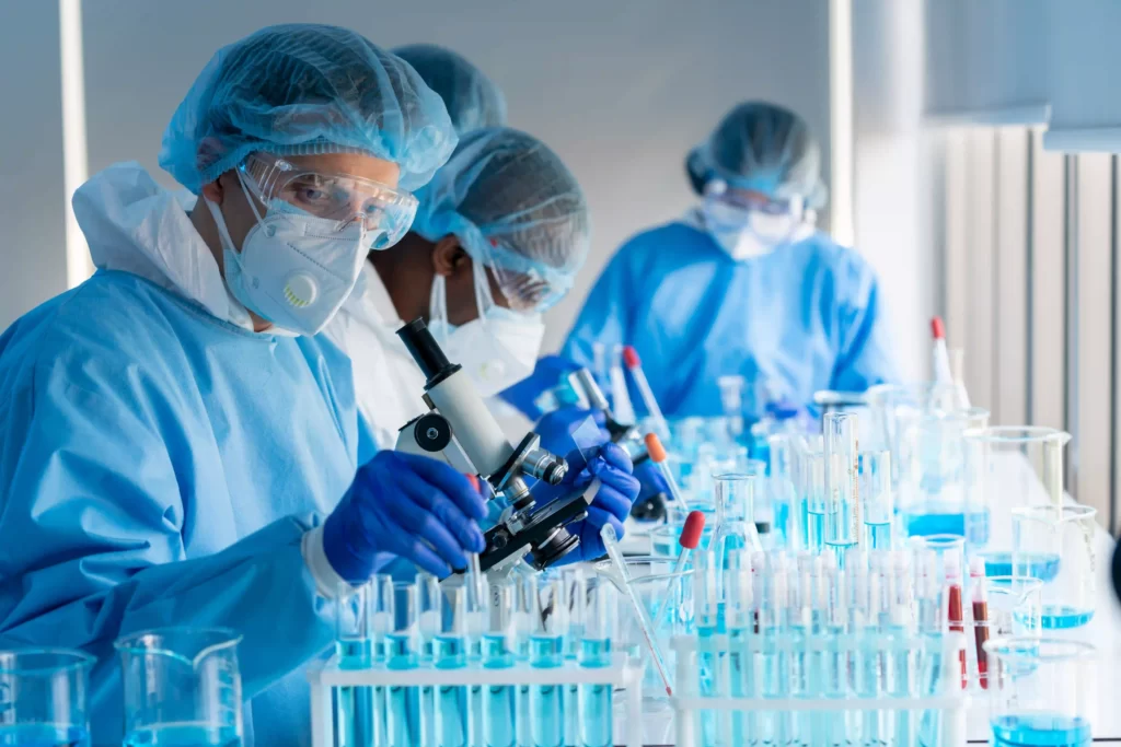 Life-science scientists in a hazardous laboratory setting, dressed in protective laboratory outfits and safety glasses, working with microscope and liquid substances in various labware.