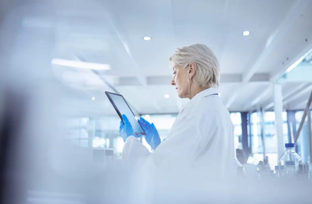 A female life-science scientist a laboratory setting, dressed in a medical gown, studying information on a tablet.