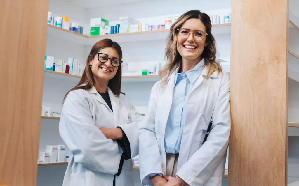 Two female life-science scientists or pharmacists in medical gowns, standing in a small room. Small boxes on shelves containing medical products.