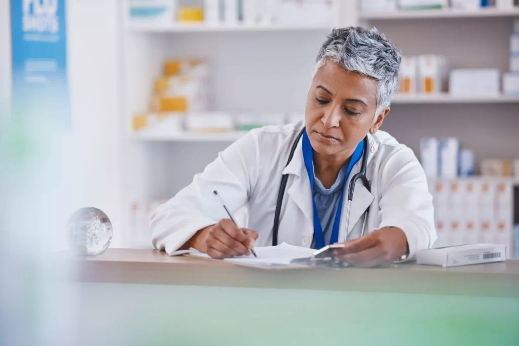 A female life-science scientist or medical doctor, in a medical gown, sitting at a desk using a pencil writing in a paper document. Small boxes on shelves containing medical products.