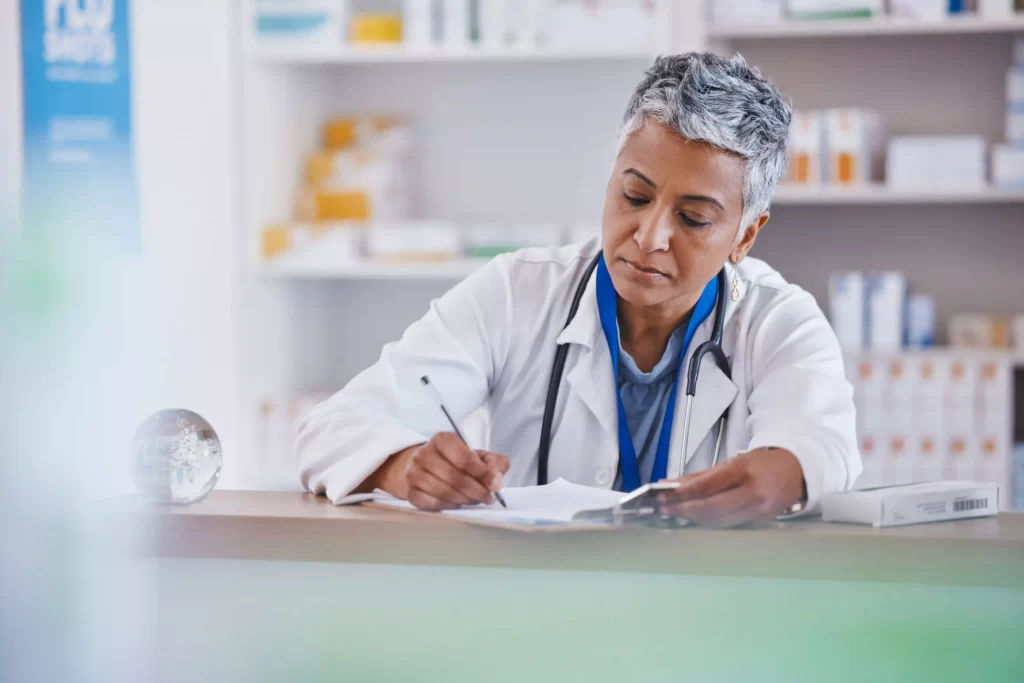 En kvinnlig forskare inom biovetenskap eller läkare, iförd läkarrock, sitter vid ett skrivbord med en penna och skriver i ett pappersdokument. Små lådor på hyllor som innehåller medicinska produkter.