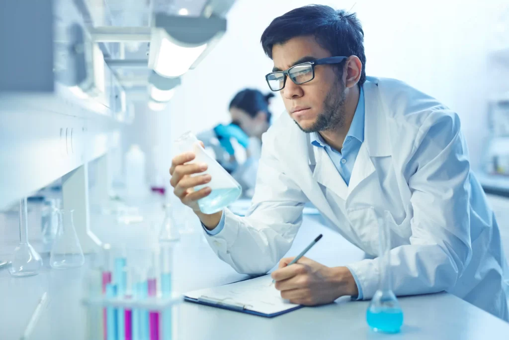 A male life-science scientists in a laboratory setting, dressed in a medical gown studying a flask labware and taking notes.