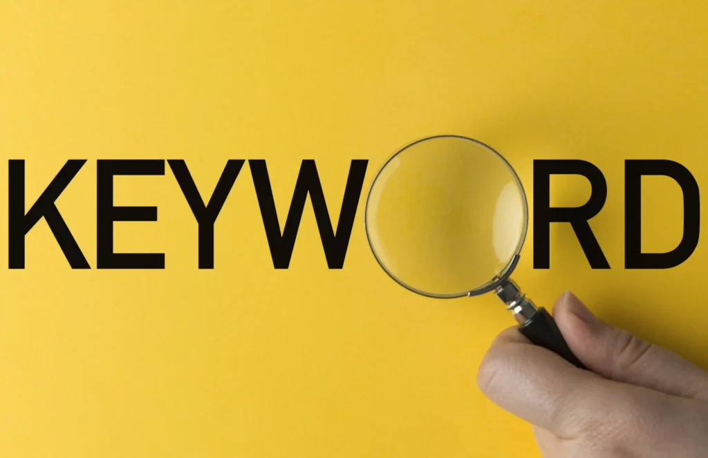 Hand holding a magnifying glass over the word "KEYWORD" on a yellow background, symbolizing keyword search and SEO focus.
