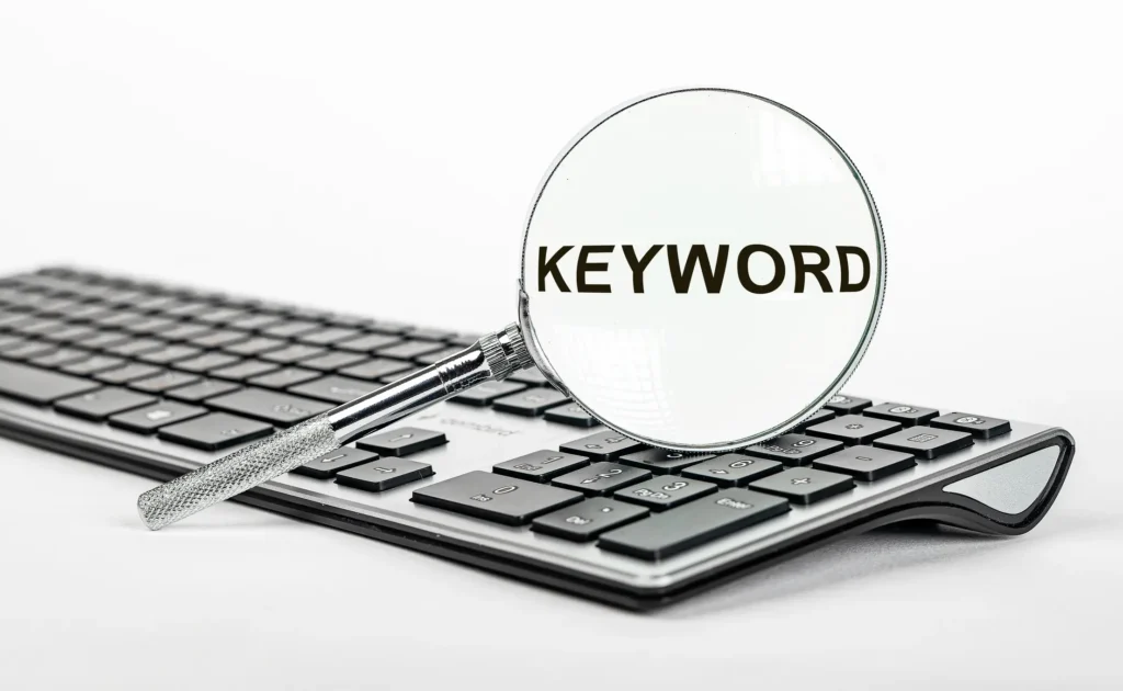 A magnifying glass placed over a computer keyboard highlights the word “keyword,” symbolizing detailed research, analysis, and focused SEO optimisation.